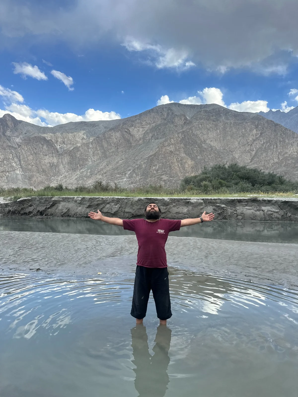 Abdullah standing in clear shallow water with arms open to the mountains